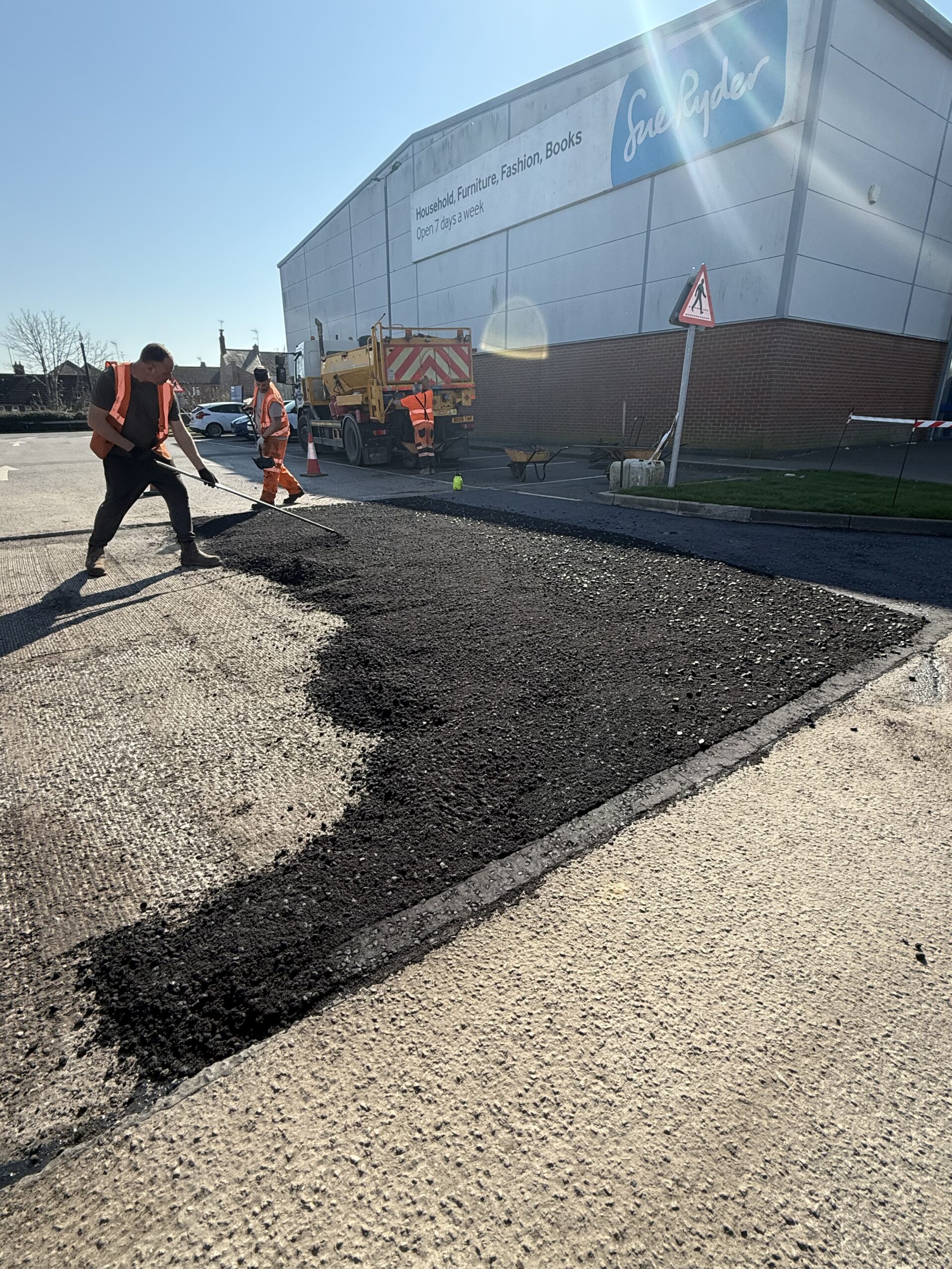Operative spraying bitumen emulsion tack coat onto prepared car park surface before tarmac laying, with steam visible from the hot bond coat application