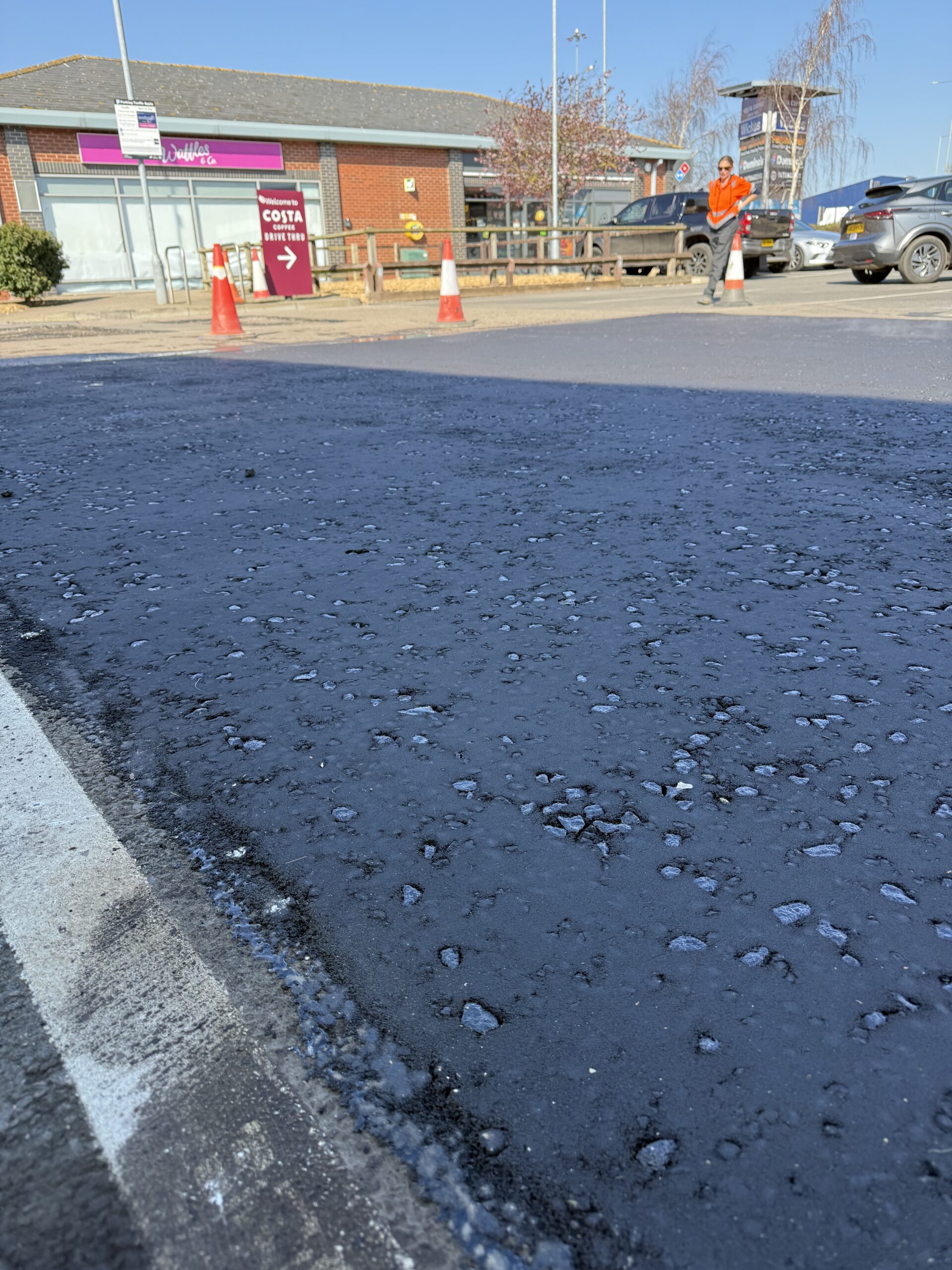 Hot-mix asphalt being discharged from a tarmac wagon into a wheelbarrow on a commercial car park resurfacing project, with steam rising from the freshly delivered material